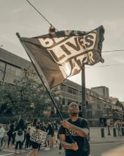 a Black man waves a Black Lives Matter flag in the middle of an intersection while a group of protesters holding signs gathers behind him