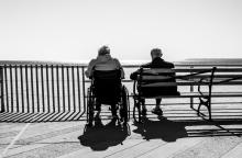 Two elderly adults sitting on a bench and wheel chair, looking out over the water