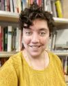A headshot of Professor Rosalind Kichler standing in front of bookshelves. Professor Kichler has short, brown curly hair and green eyes